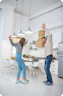 White married couple in their mid-thirties wearing casual attire dancing and celebrating in their all-white kitchen after learning they have grown their equity.