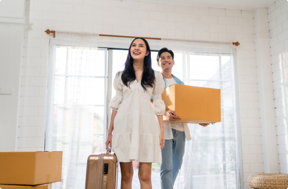 Young asian couple enters their new home for the first time, smiling, carrying boxes and a suitcase.