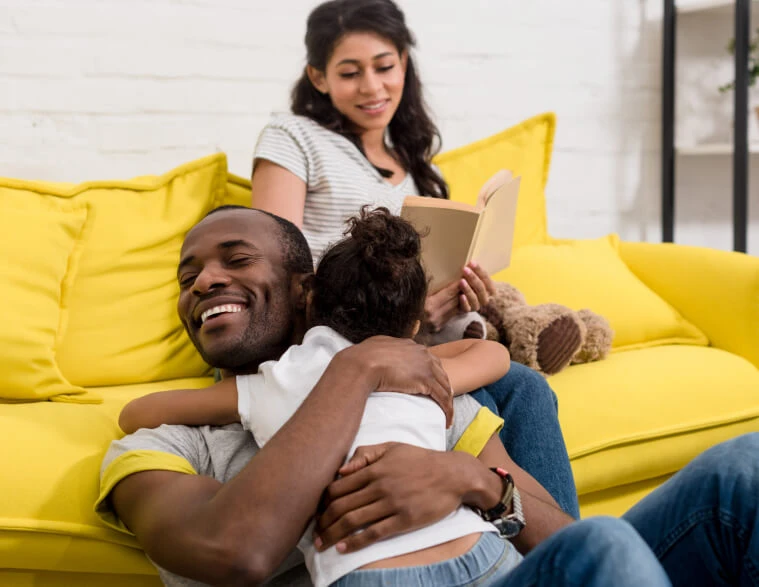 Mixed-raced family celebrating the funding of their first home. Young daughter hugging her father as the Mom admires from a short distance on the couch.