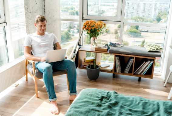 Young white male with an arm tattoo sitting on a chair smiling in his window-filled bedroom, with his laptop reading up about cash-out refinancing.