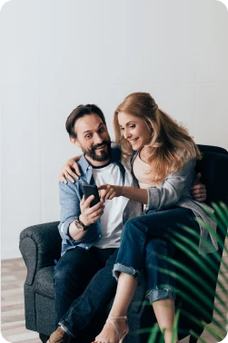 A young couple sitting on a chair together playing around with the Golden Coast Finance QuickQualify tool on a cell phone, both of them smiling down at the phone.